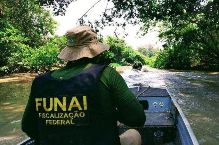 Visão por trás de um agente da Fundação Nacional dos Povos Indígenas (Funai) vestindo um colete de Fiscalização Federal e chapéu, sentado em um pequeno barco a motor navegando em um rio estreito cercado por mata densa.