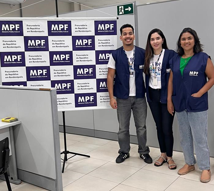 três servidores - um homem e duas mulheres - aparecem ao lado de um banner do MPF, onde a logo do órgão aparece repetida várias, uma do lado do outro, cobrindo todo o espaço do banner