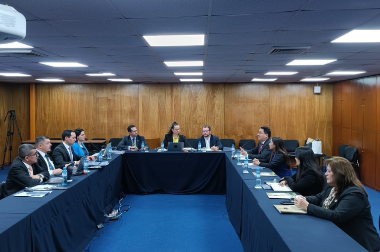 Foto mostra cinco mulheres e seis homens sentados em torno de mesa em formato de U em sala de reunião. A mesa está coberta com toalha azul. O tapete da sala é azul e as paredes são revestidas de madeira.