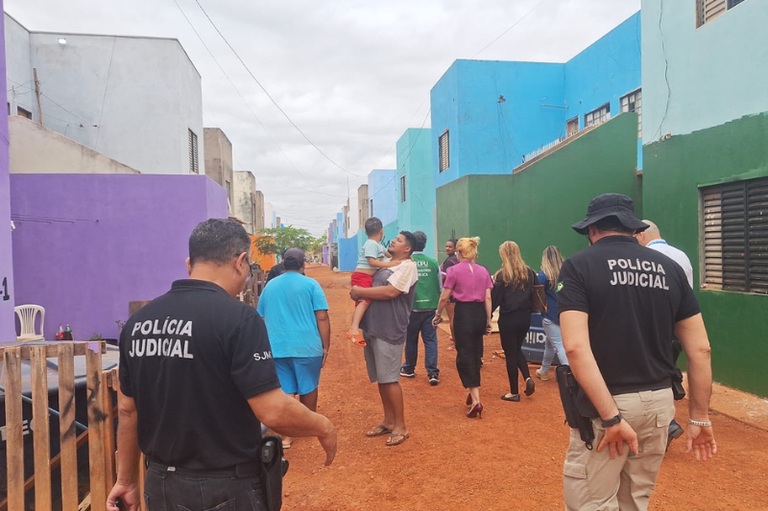 Agentes da Polícia Judicial e uma equipe de pessoas (civis) caminham por uma rua de terra vermelha em uma área residencial caracterizada por casas geminadas coloridas (azul, verde, roxo e branco). No centro, uma pessoa carrega uma criança ao colo, e os agentes da Polícia Judicial estão em primeiro plano.