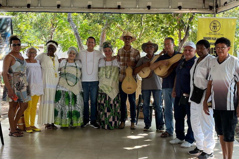 Fotografia do procurador-chefe do MPF em Mato Grosso, Ricardo Pael Ardenghi, e mais seis homens, três dos quais seguram instrumentos musicais parecidos com violões, e cinco mulheres, sob uma tenda, no território Ilê Asé Idasilè Opô Oluayê, no município de Santo Antônio do Leverger (MT). A foto foi registrada no dia 26/04/2025, durante visita do MPF ao Projeto Baobás pelo Brasil, a convite do Movimento Negro Unificado
