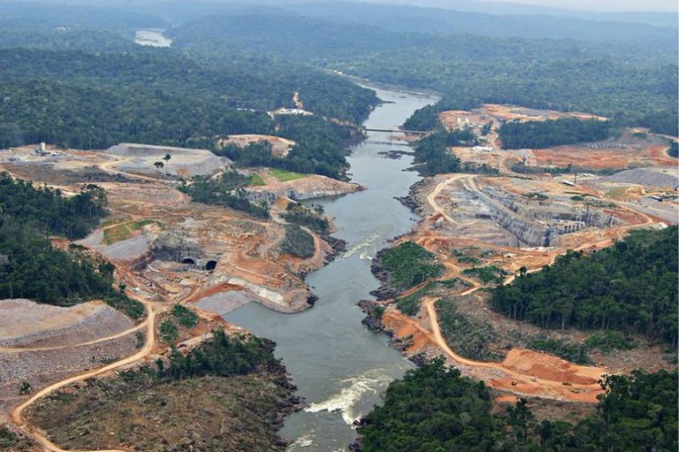 Vista aérea das obras de construção de uma usina hidrelétrica ao longo de um rio, cortando uma vasta área de floresta tropical.