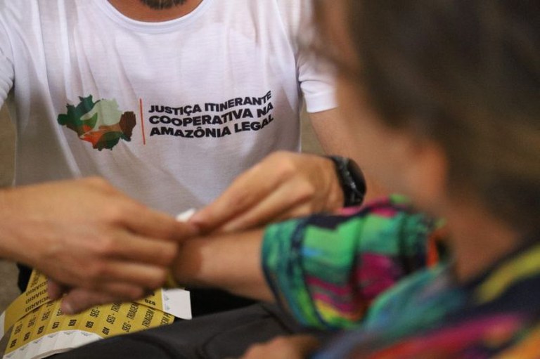 Homem com camiseta branca escrita "Justiça Itinerante Cooperativa na Amazônia Legal" coloca pulseira amarela em braço de pessoa