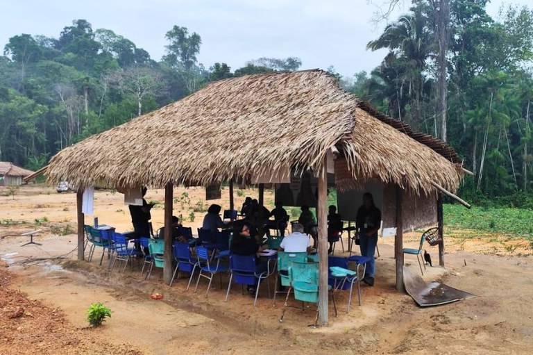 Foto mostra indígenas em cadeiras escolares debaixo de uma estrutura improvisada de palha