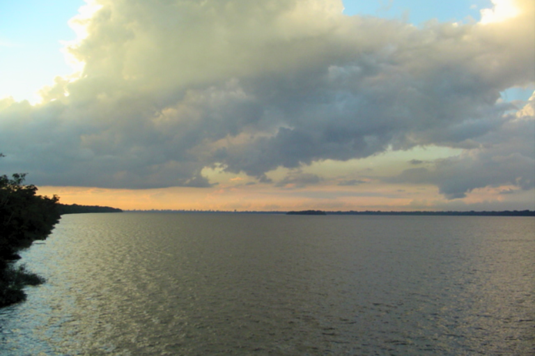 Foto mostra um grande corpo de água (a baía do Guajará) com superfície levemente ondulada, ocupando a maior parte do quadro, e uma faixa de vegetação escura à esquerda. O horizonte apresenta uma linha de terra baixa ao fundo e o céu está coberto por nuvens densas e escuras, com áreas iluminadas em tons claros próximos ao pôr do sol.