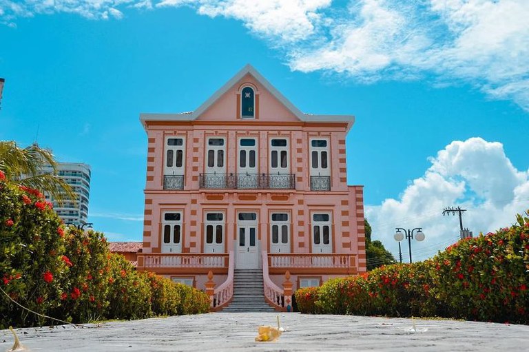 Casarão rosa com detalhes em laranja e janelas brancas, escada central, cercado por arbustos com flores vermelhas sob céu azul.