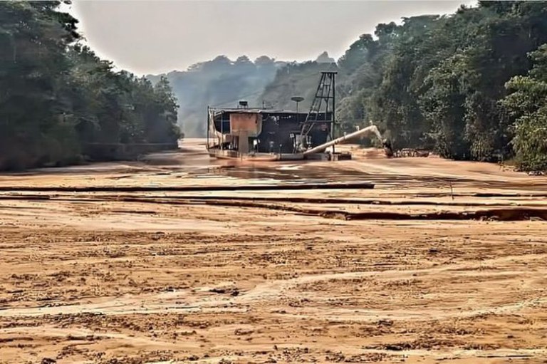 Draga de mineração em leito de rio seco e barrento, cercada por mata densa sob céu claro e ensolarado.