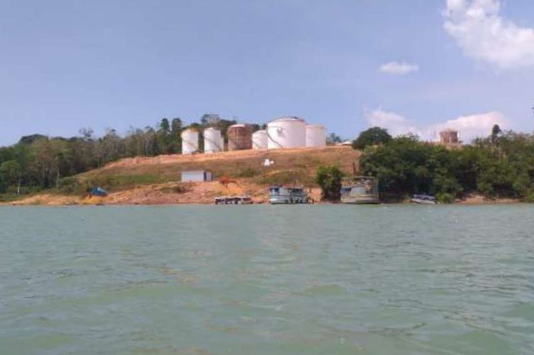 Foto em formato retangular de um porto em construção às margens do rio Tapajós. Vários tanques de armazenamento brancos, de grande porte, estão instalados em uma elevação na margem. Próximo à água, há algumas estruturas e embarcações menores.