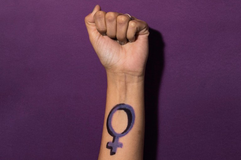 Um braço erguido com punho fechado e o símbolo do feminino pintado em roxo no pulso, sobre fundo roxo escuro.