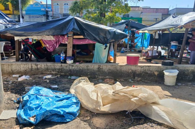 Foto de acampamento improvisado com barracas feitas de lonas plásticas pretas e coloridas, sustentadas por madeira, em área do centro de Belém, no Pará. No chão, há lonas rasgadas, lixo espalhado e baldes plásticos. Ao fundo, pessoas aparecem em meio às barracas, com casas e comércios visíveis ao longe.