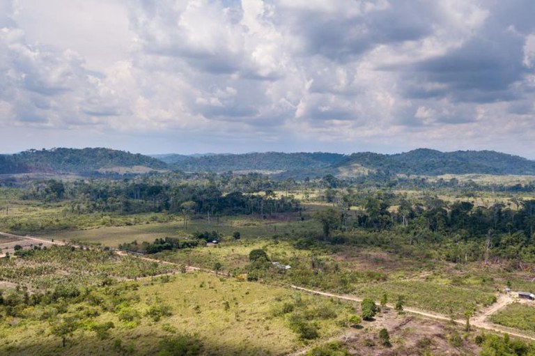Vista aérea de paisagem rural com colinas ao fundo sob céu nublado. Em primeiro plano, áreas abertas, estradas de terra e árvores.
