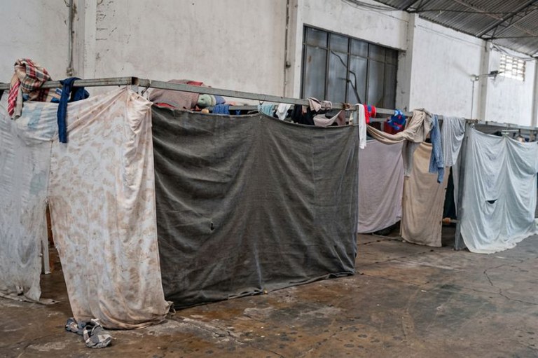 Galpão com estruturas metálicas onde cobertores e panos estão pendurados, formando divisórias. Roupas apoiadas e sandálias no chão.