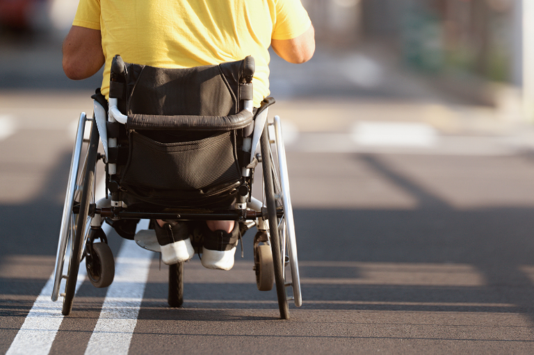 #ParaTodosVerem: Pessoa de costas em cadeira de rodas sobre faixa branca no asfalto. Veste camiseta amarela e tênis pretos. Fundo desfocado com luz solar.