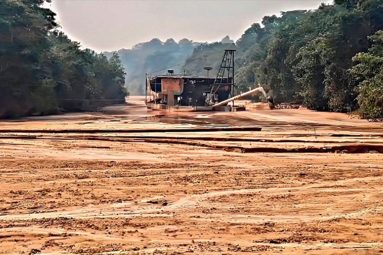 Foto retangular do leito de um rio seco e onde há uma draga de garimpo encalhada. As margens do rio são cobertas por floresta densa.