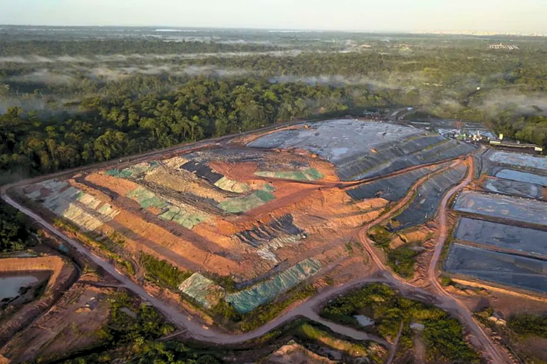Vista aérea do aterro sanitário de Marituba, com áreas de terra e lonas plásticas escuras e esverdeadas, cercado por densa floresta com névoa ao fundo e tanques ao lado.