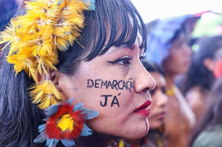 Mulher indígena de perfil com pintura no rosto "Demarcação já", cocar e brinco de penas coloridas. Ao fundo, outras pessoas em desfoque.