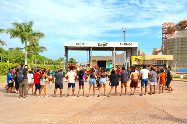 Pessoas, algumas com cocares, em roda de mãos dadas diante de uma portaria com letreiro escrito Cargill.