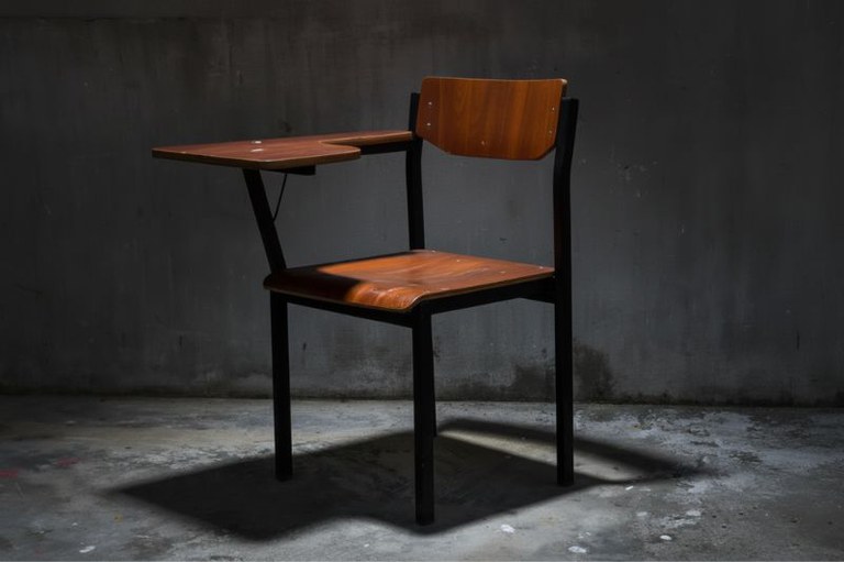 Foto de uma cadeira escolar vazia com mesa acoplada, de estrutura preta e assento e encosto de madeira, em um ambiente escuro.