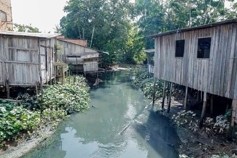 Rio com águas escuras ladeado por casas de madeira suspensas sobre palafitas e vegetação verde ao fundo.