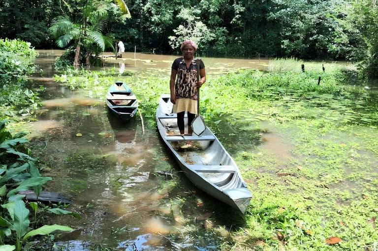Mulher de pé em uma canoa segura um remo em um rio cercado por vegetação densa. Ao lado, há outra canoa vazia na água.