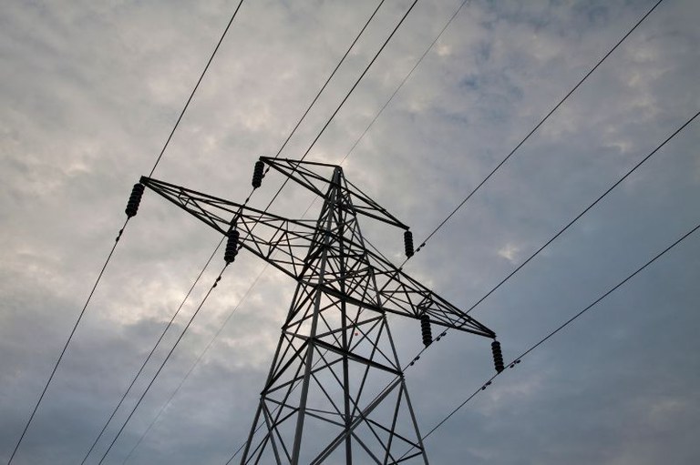 Torre de transmissão de energia metálica em contraluz sob céu nublado com vários cabos elétricos estendidos na diagonal