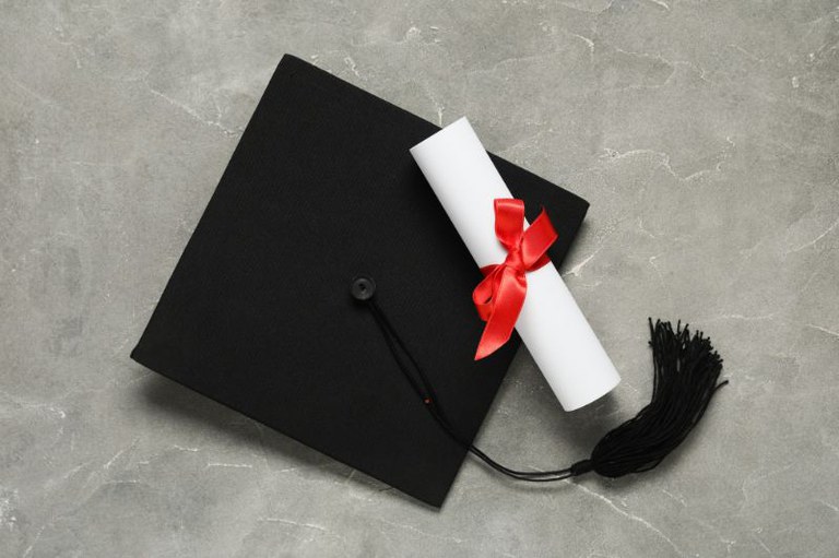 Vista superior de um capelo preto e um diploma enrolado com fita vermelha sobre uma superfície cinza marmorizada.