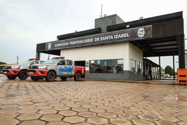 Foto da fachada do Complexo Penitenciário de Santa Izabel, com duas viaturas da Polícia Militar estacionadas sobre o piso de paralelepípedos.