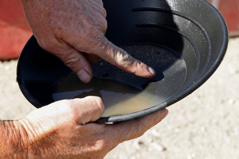 Foto em close das mãos de uma pessoa que segura uma bateia de garimpo preta. Um dedo aponta para o sedimento escuro e a água no fundo da bateia.