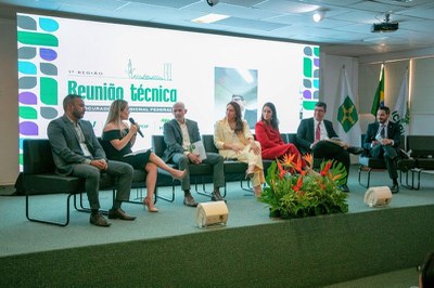Foto de sete pessoas sentadas em um palco diante de uma tela com a frase Reunião Técnica e arranjo de flores tropicais à frente.
