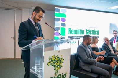 Homem de terno assina documento em púlpito transparente do ICMbio. Ao fundo, tela exibe o texto Reunião técnica e há pessoas sentadas no palco.