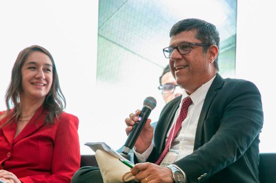Homem sorridente de terno e óculos fala ao microfone. Ao seu lado, mulher sorri usando terno vermelho.