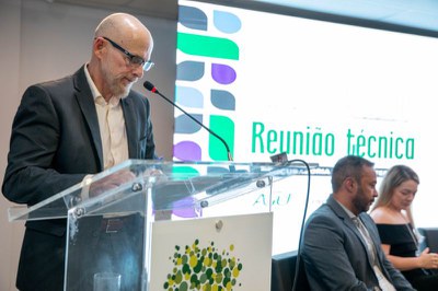 Homem calvo de barba grisalha e terno lê no púlpito. Ao fundo, a tela exibe Reunião técnica e duas pessoas estão sentadas no palco.