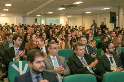 Auditório lotado com pessoas vestidas formalmente em poltronas verdes assistindo a um evento e aplaudindo.