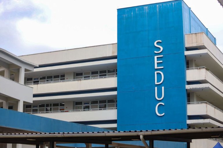 Foto da fachada de um prédio de vários andares, com paredes brancas e detalhes em azul, incluindo uma coluna onde está escrito Seduc em letras brancas na vertical.
