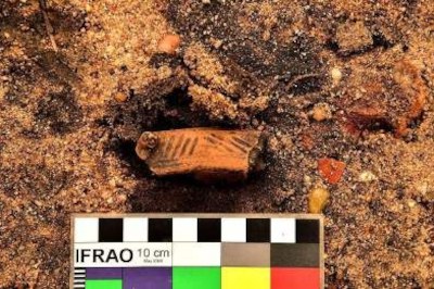 Foto em close-up de um achado arqueológico: um pequeno fragmento de cerâmica cilíndrico, de cor terracota com padrões de linhas diagonais pretas, repousa sobre terra arenosa e escura. Na parte inferior da imagem, há uma régua de escala de 10 cm, para referência. A régua cita a sigla Ifrao, sigla em inglês da Federação Internacional de Organizações de Arte Rupestre.