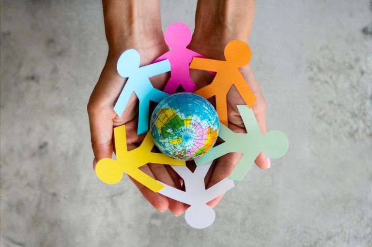 Duas mãos seguram recortes de papel coloridos em forma de pessoas em círculo ao redor de um pequeno globo terrestre