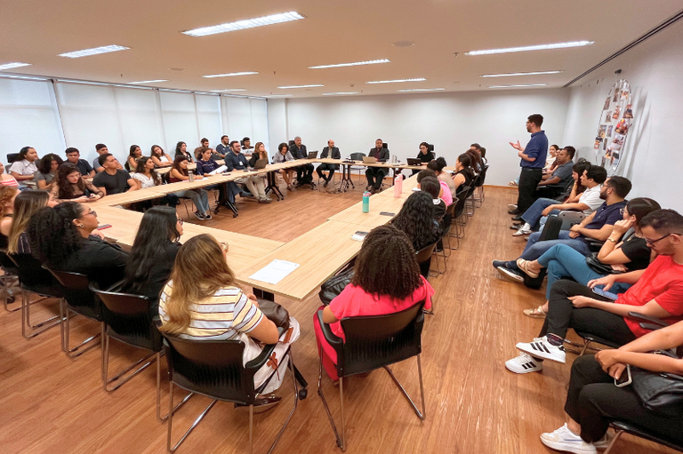 Foto em plano aberto de uma reunião em sala clara com mesas em formato de "U". Dezenas de pessoas estão sentadas ao redor das mesas e em fileiras laterais enquanto um homem fala de pé à direita