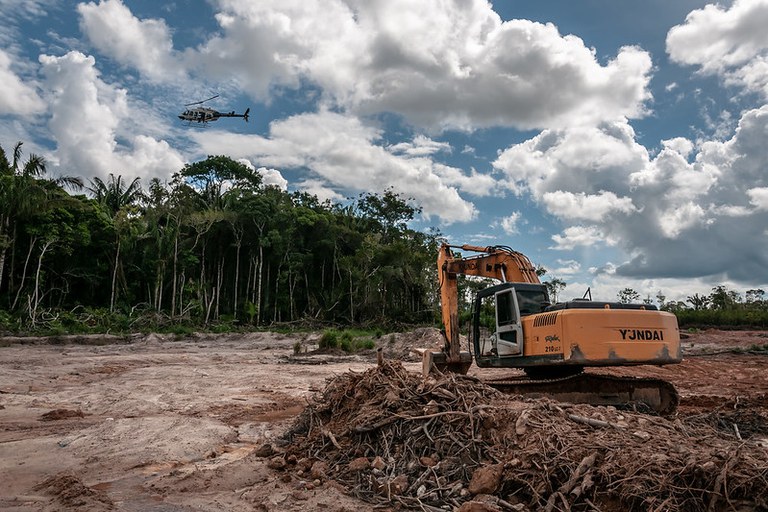 Foto de escavadeira amarela parada em uma área de terra desmatada em frente a uma floresta densa, enquanto um helicóptero voa no céu acima.