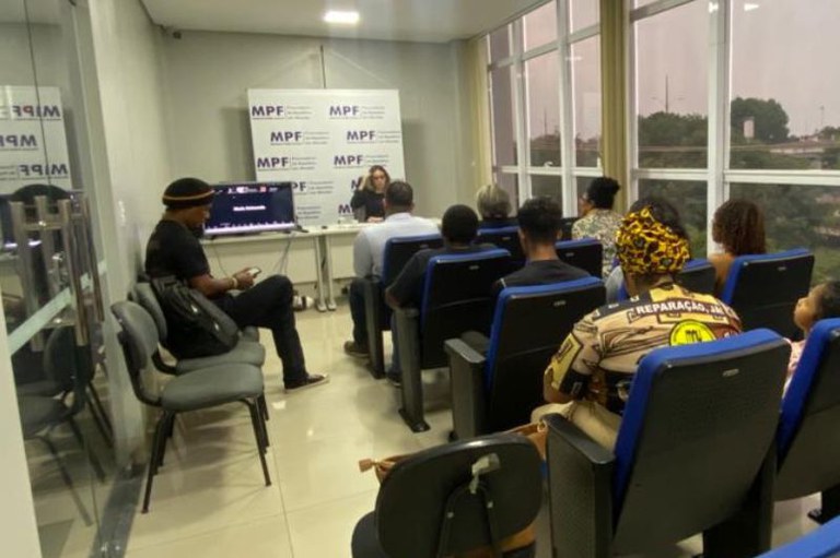 Pessoas assistem a um evento em uma sala com janelas, cadeiras azuis e cinzas, e um banner do MPF ao fundo.