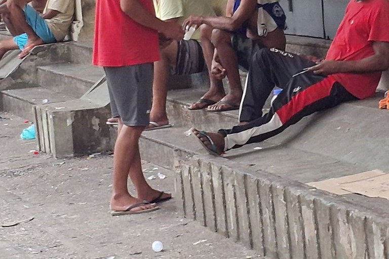 Pessoas sentadas e em pé em escadaria de concreto com lixo espalhado pelo chão e degraus. Predominam roupas casuais e chinelos de dedo.