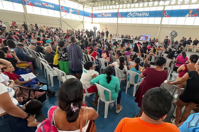 Um grande público de costas, sentado em cadeiras brancas num ginásio, assiste a um painel de pessoas e uma tela ao fundo, sob faixas com a palavra "Castanhal"