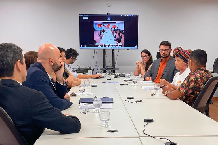 Homens e mulheres sentados ao redor de uma mesa branca de reunião com uma tela ao fundo que mostra uma videochamada.