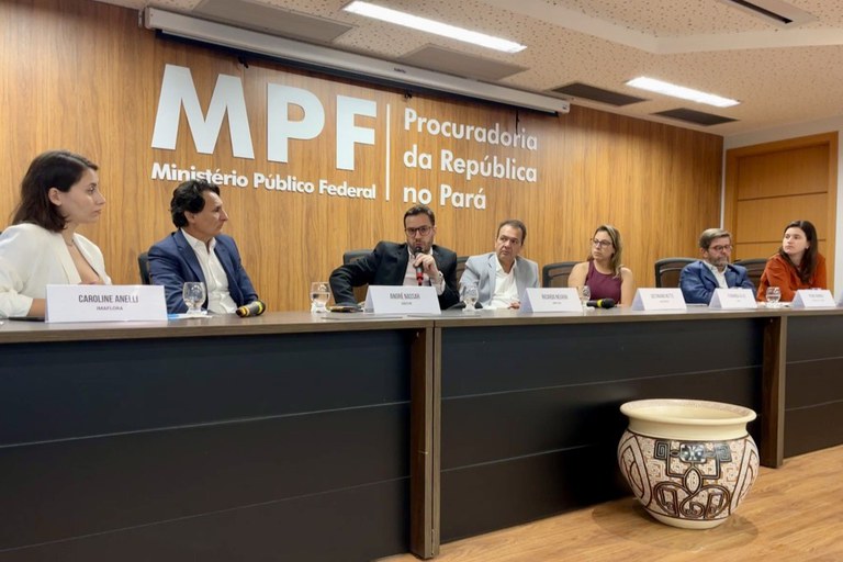 Sete pessoas em mesa de painel em auditório com a logo do MPF ao fundo. Um homem fala ao microfone. À frente, um vaso de cerâmica marajoara no chão.