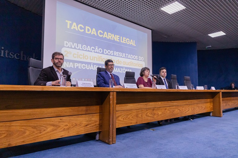 Foto de quatro pessoas, dois homens e uma mulher, atrás de uma mesa de madeira no palco de um auditório. Ao fundo, está escrito TAC da Carne Legal - Divulgação dos Resultados do 2º ciclo unificado de auditorias na pecuária da Amazônia
