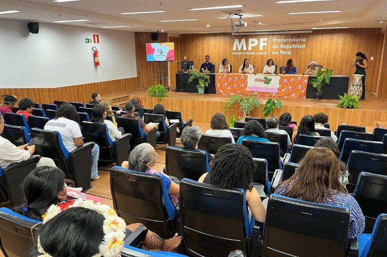 Pessoas sentadas em poltronas azuis assistem a uma mesa com sete pessoas sentadas à frente de uma tela com a sigla CPDDH. Ao fundo do palco, a logo do MPF.