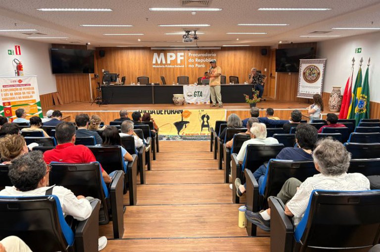 Auditório com público sentado assistindo a um homem falar ao microfone no palco, sob a sigla MPF em parede de madeira ao fundo.