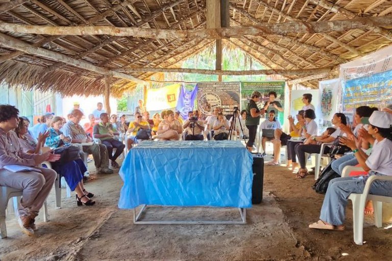 Foto de um grupo de pessoas reunidas sob uma estrutura de telhado de palha. Há uma mesa retangular coberta com um tecido azul no centro, em frente a um grupo de pessoas sentadas em cadeiras, algumas aplaudindo. Ao fundo, há banners e cartazes pendurados. O chão é de terra batida.