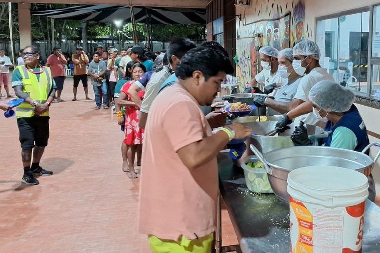 Foto de pessoas em uma fila em um local coberto recebendo comida servida por voluntários em uma bancada com grandes panelas.