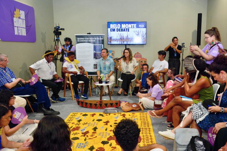Debate com indígenas e público sentado em círculo no chão e cadeiras, banner com texto e TV exibindo o tema.