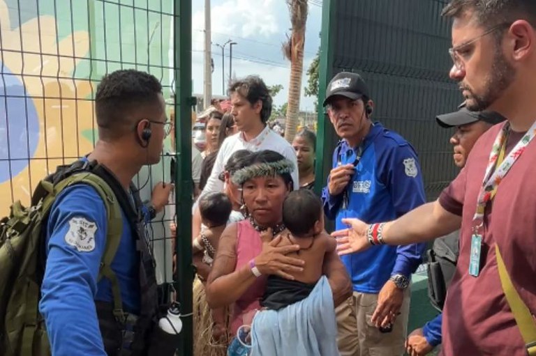 Foto de uma mulher indígena com um cocar e um bebê no colo, em meio a um grupo de pessoas. Ela é recebida na Zona Verde da COP30 pelo procurador regional dos Direitos do Cidadão no Pará, Sadi Machado, homem com barba e óculos.